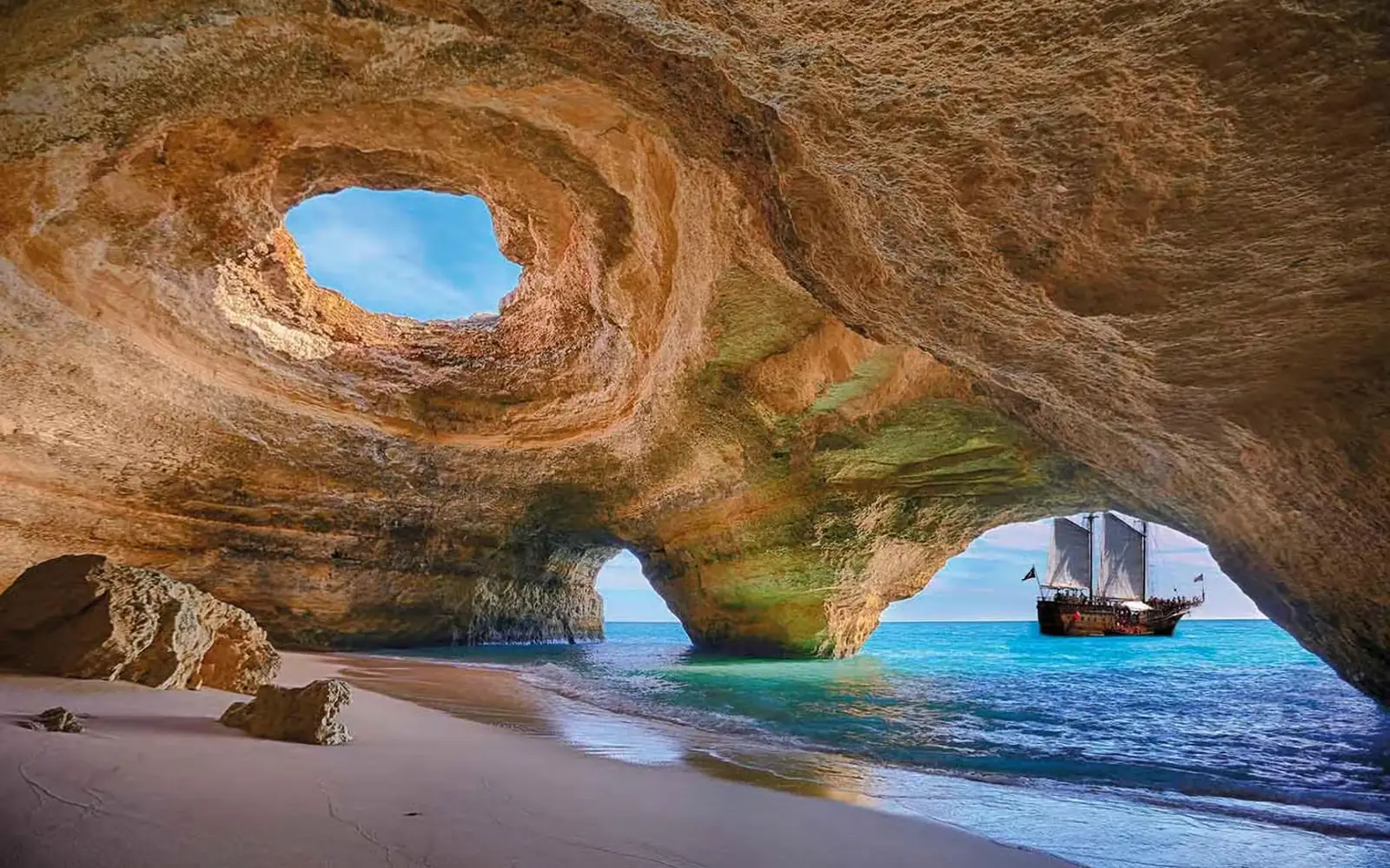 Inside the Benagil cave: the round opening in the domed roof, the beach below, and a tall ship on the Atlantic through the arch